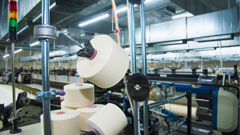 Interior of a textile factory with large spools of white thread on machinery, and overhead ventilation ducts.
