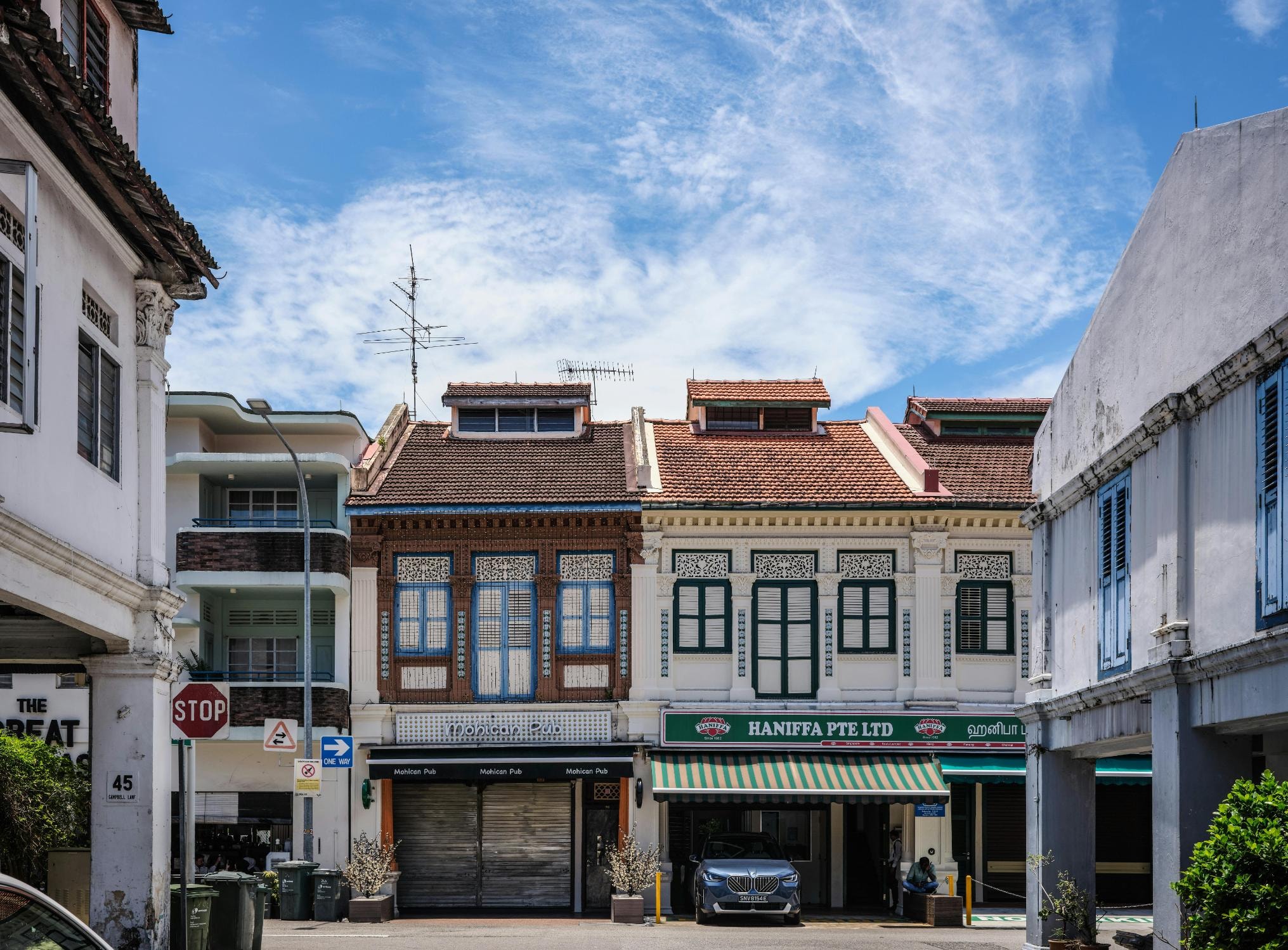 Under a bright blue sky scattered with wispy clouds, a row of traditional shophouses showcases intricate architectural details, including a white facade with green shutters for "Haniffa Pte Ltd" and an adjacent building housing "Mohican Pub".