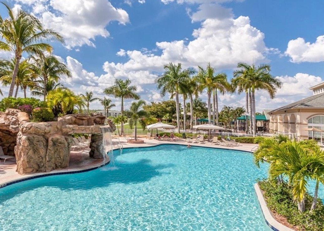 The image depicts a vibrant community pool area at the Legends Golf and Country Club, where residents are engaging in social activities and enjoying the recreational facilities. Surrounding the pool are lush landscapes and beautiful water features, creating a picturesque setting for relaxation and community interaction.