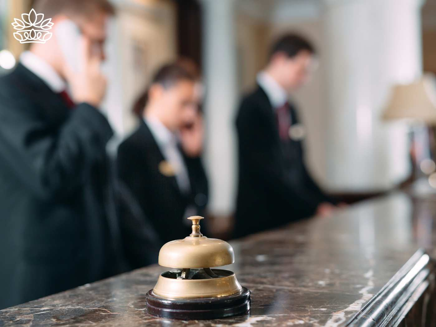 Focused scene at a luxury hotel reception with staff members actively engaged in service. One concierge speaks on the phone, another assists a guest, and a third attentively manages paperwork. In the foreground, a classic brass service bell sits on the marble countertop, ready to summon assistance. Fabulous Flowers and Gifts for Guest House and Hotel Flowers
