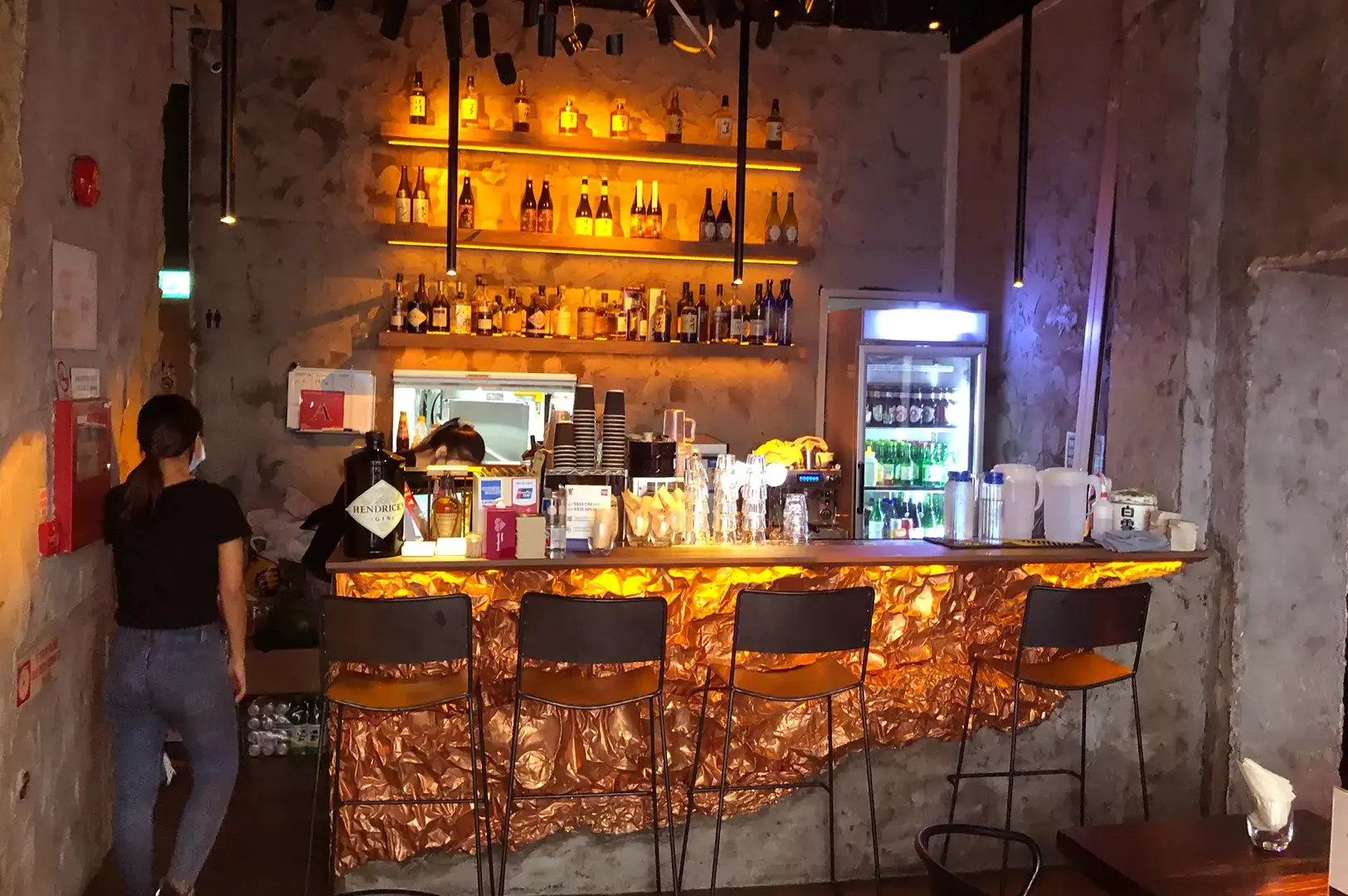 TAAN IZA-BAR interior with a warmly lit bottle shelf, copper-fronted counter, stools, drinks refrigerator, and a staff member.