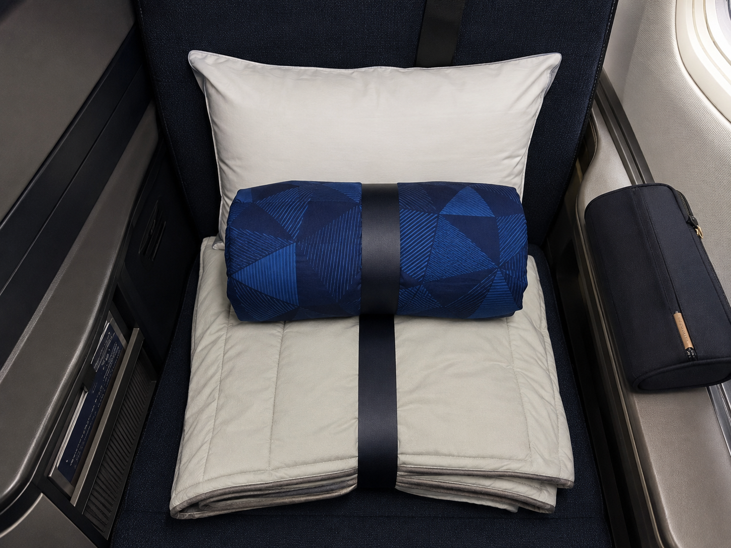 Business class seat with a white pillow, folded light-grey blanket, blue patterned bedding and a navy amenity pouch beside the window.