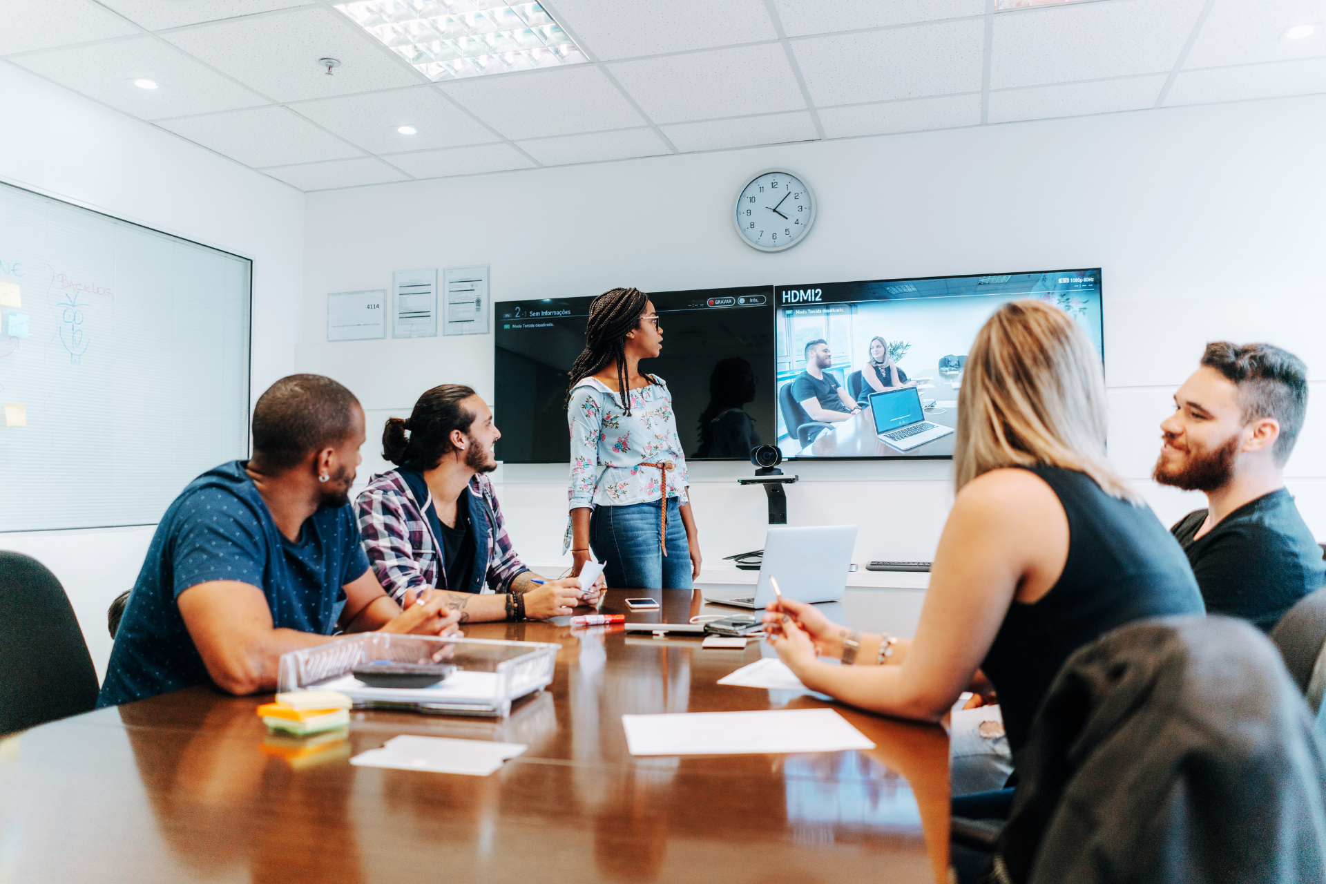 A woman presenting to a team in a conference room with dual screens showing a video call with remote colleagues.