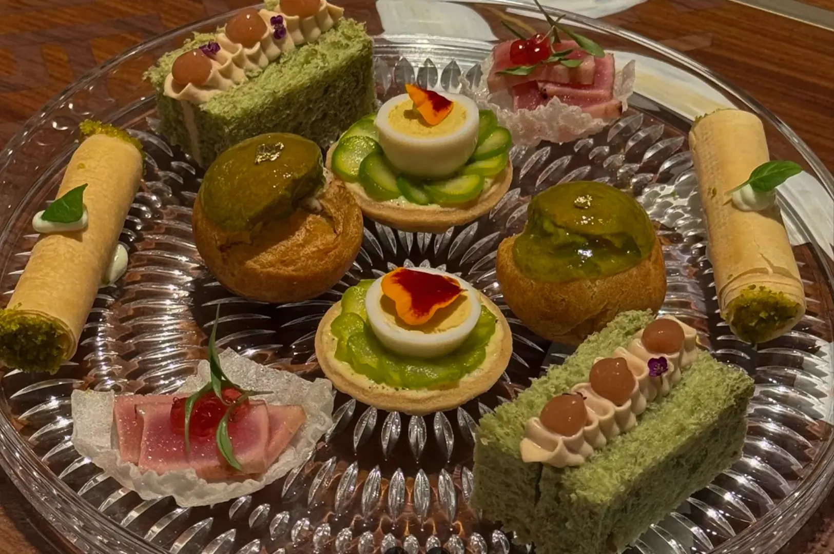 A detailed top-down view shows an assortment of gourmet hors d'oeuvres arranged symmetrically on a clear, textured glass platter resting on a wooden surface. The selection includes two rectangular green sponge cakes topped with piped cream and jelly drops, two round tarts filled with green asparagus slices and a halved soft-boiled quail egg garnished with an edible petal, two golden choux pastries coated in a shiny green glaze with gold leaf accents, two tightly rolled crispy pastry cylinders filled with green pistachio cream, and two delicate pieces of seared tuna topped with red roe and a microgreen leaf.