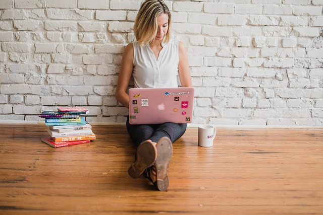 A woman sitting on a wooden floor; laptop sitting on her lap with her leg cross. With a pile of books on one side and a coffee mug on the other.