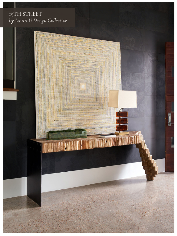 A dramatic entryway with a dark textured wall, a large square geometric artwork in soft gold tones, and a sculptural console table with wood and black metal detailing. A table lamp sits on one side of the console