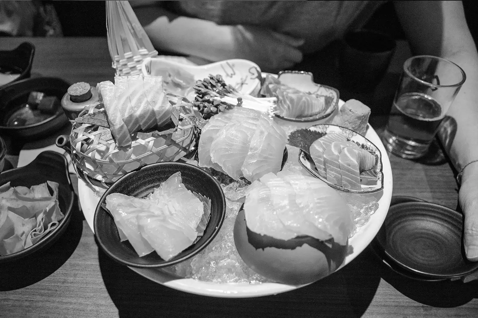 Assorted sashimi and sushi arranged on a large platter with ice, garnishes, dipping sauces, and drinks on a restaurant table.