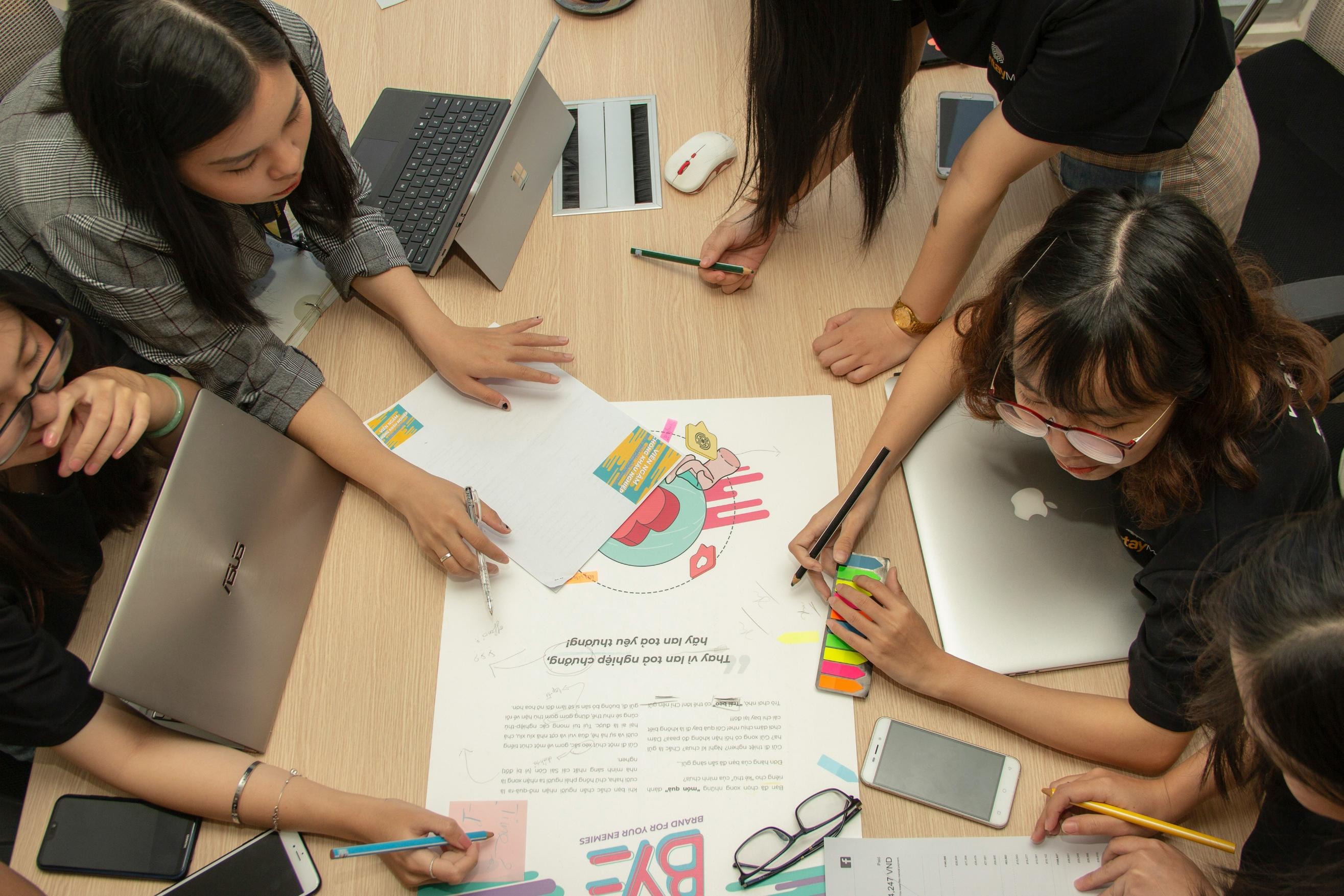 This high-angle shot depicts a group of individuals collaborating around a wooden table, utilizing laptops, smartphones, and stationery to brainstorm on a large sheet of paper. The scene conveys a focused and creative atmosphere as they actively write notes, draw diagrams, and discuss ideas together.