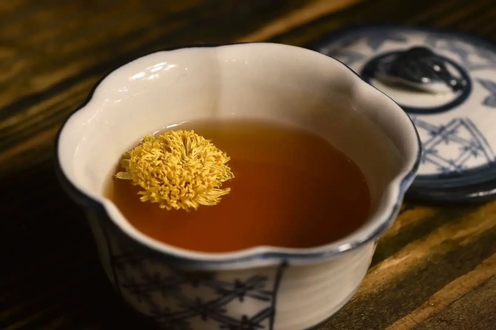 A close-up shot of an elegant white ceramic teacup with blue grid-like patterns on its base, resting on a dark rustic wooden table. A single, large golden-yellow chrysanthemum flower blooms beautifully as it floats on the surface of the amber-colored herbal tea inside the cup. In the blurred background, the matching patterned lid of the teacup lies slightly askew on the wooden surface.
