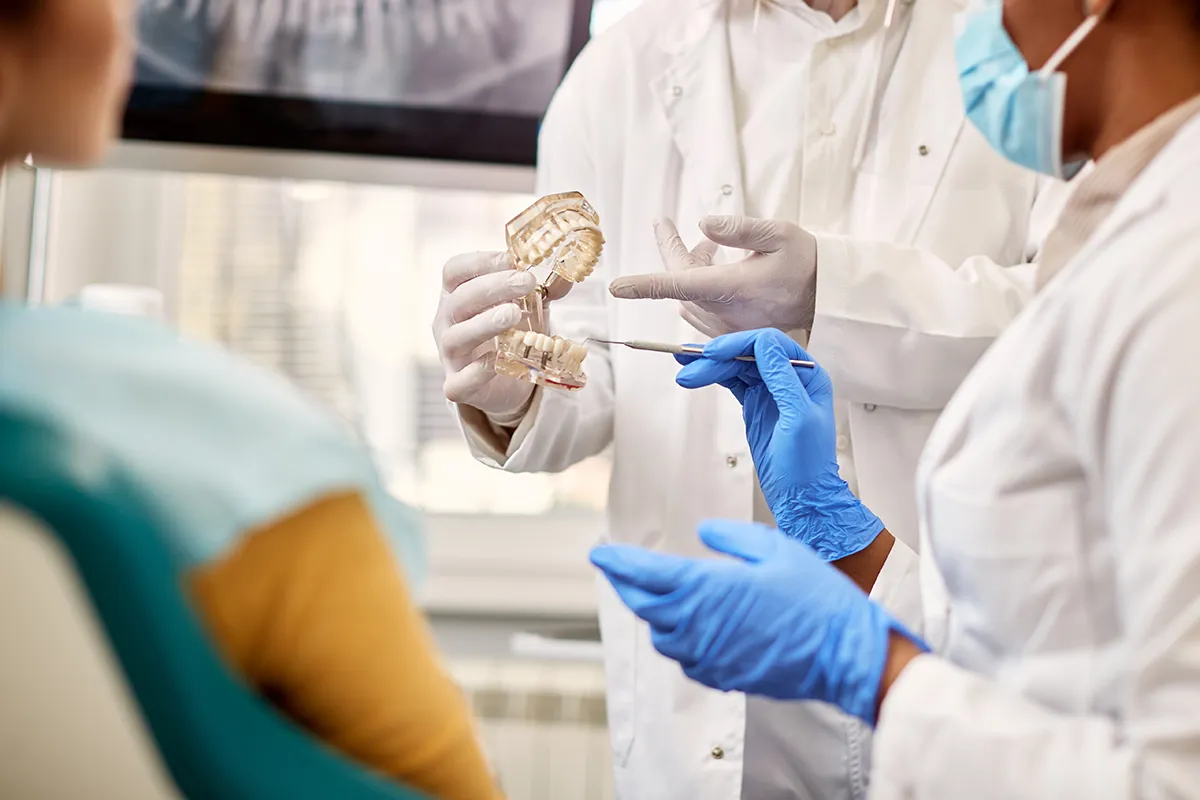Dental professionals in white coats examining a transparent dental model, with one wearing blue gloves and holding a dental tool during patient consultation. Retry