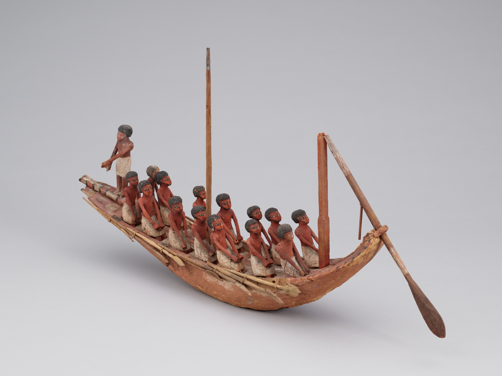A painted wooden boat with 15 wooden figures, all painted with white cloth around their lower halves. Fourteen figures are seated as if rowing; one figures stands facing sideways at the back of the boat.