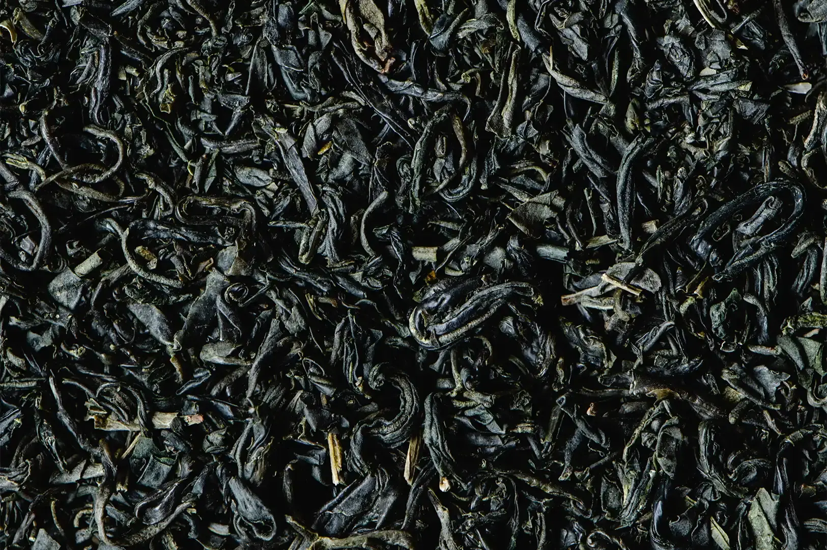 An extreme, high-contrast macro texture shot focusing on a dense pile of dry, dark-colored Lapsang Souchong tea leaves, highlighting their tightly twisted, needle-like shape and deep black and brown tones.