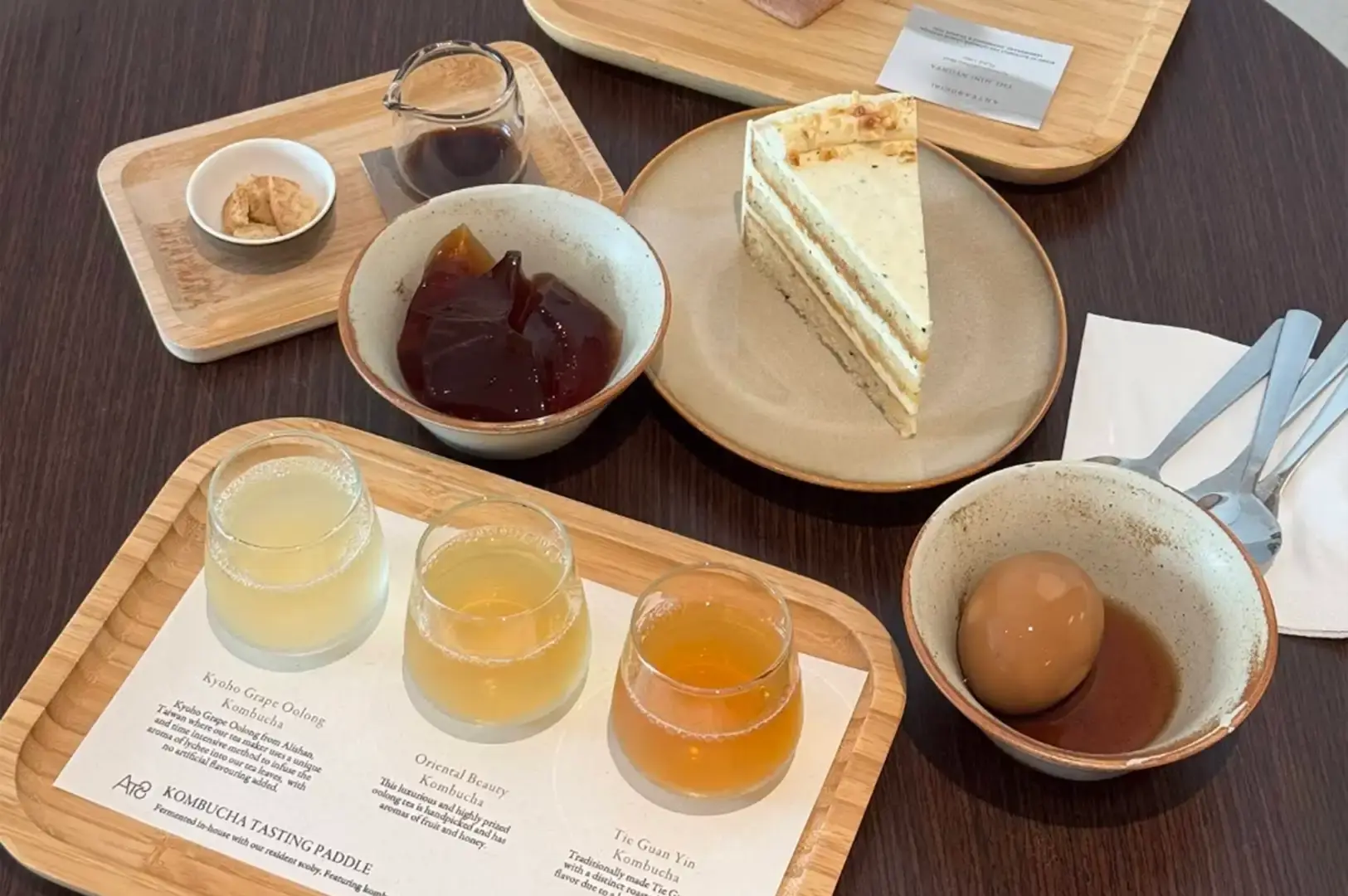 A flat-lay view displays an array of teahouse treats set on a dark wooden table. A light wooden tray in the foreground holds a kombucha tasting flight of three distinct colored brews alongside a menu card, while a ceramic bowl beside it holds a single tea-infused egg in sauce. On a plate nearby rests a slice of layered crepe cake, accompanied by a bowl of dark tea jelly and a small wooden tray with syrup and garnishes.