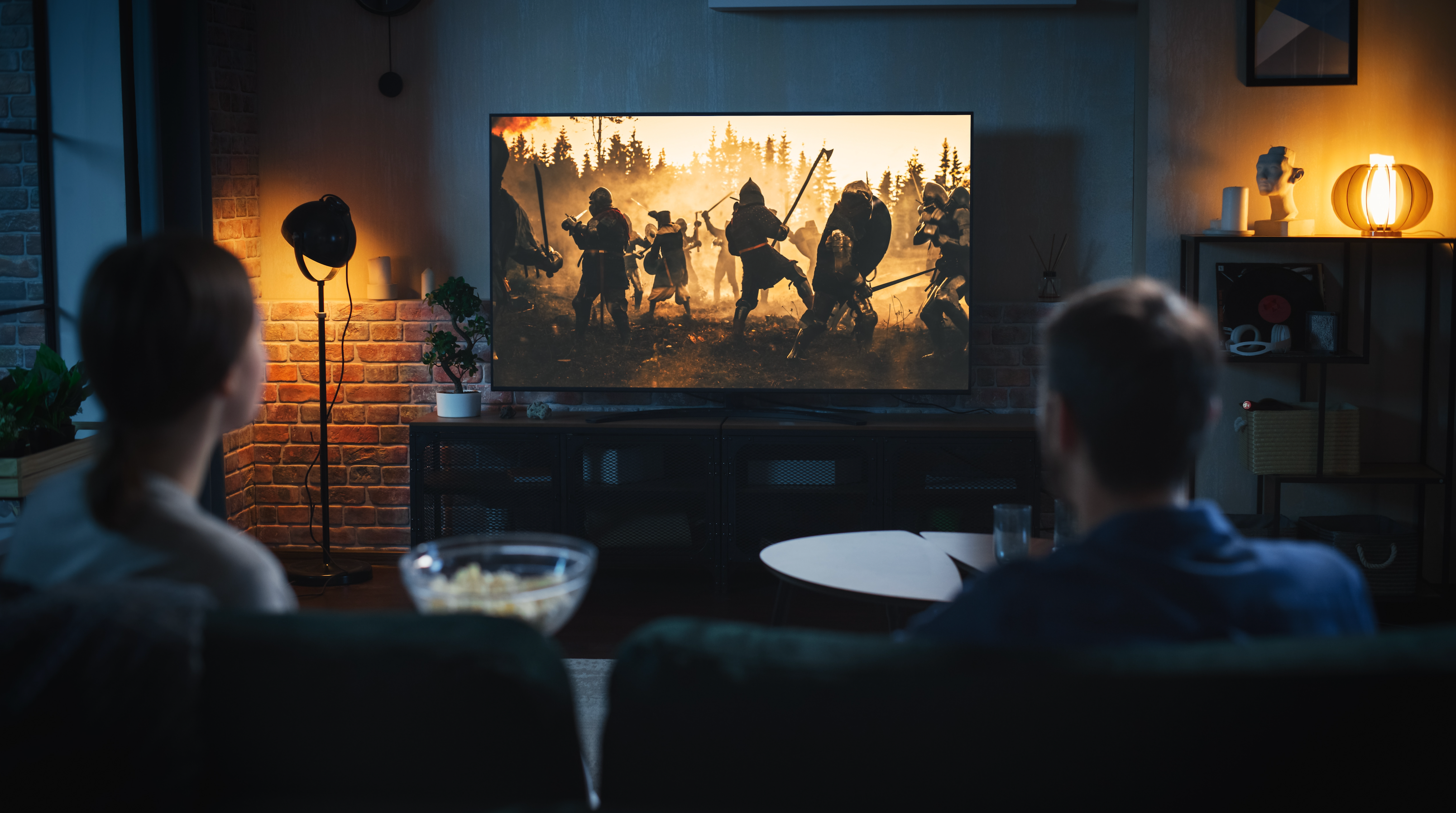 a man and woman watch an action scene on their TV as they sit on their couch