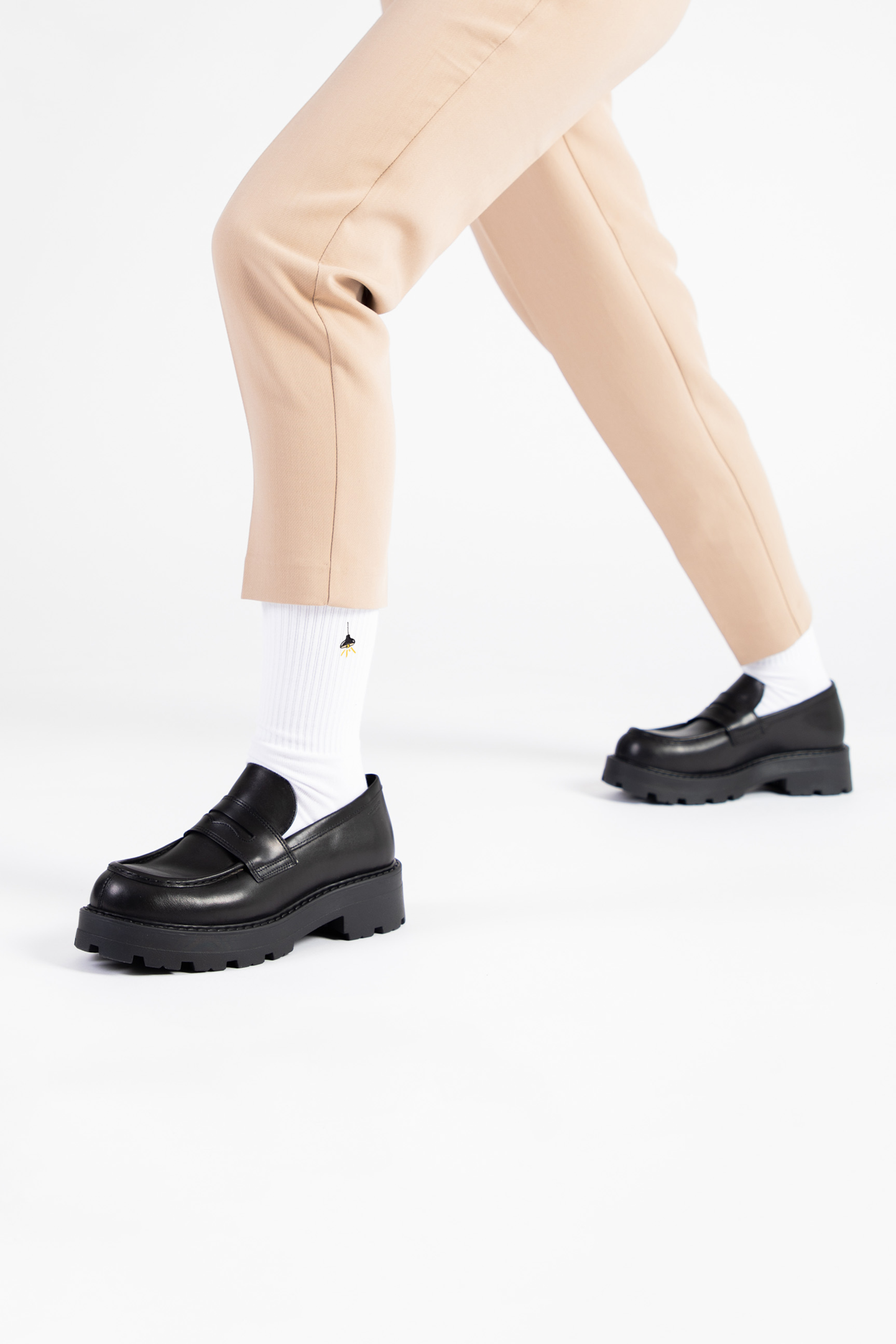 A female model wears beige tapered trousers with black leather loafers and a pair of white crew socks with a retro hanging lamp embroidered on them..