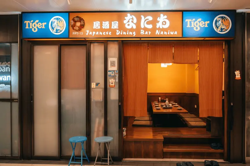 The entrance to Japanese Dining Bar Naniwa, recognizable by its orange signboard and Tiger Beer logos, offering a glimpse of traditional sunken seating areas separated by orange curtains inside.