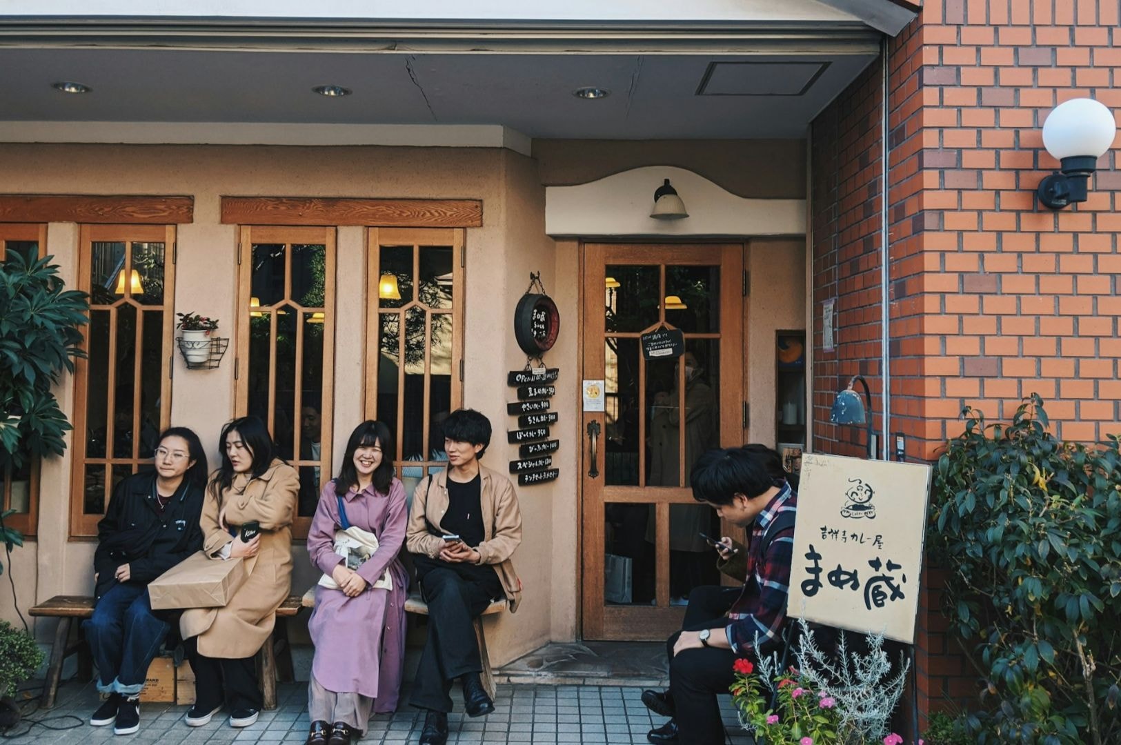 Five people sit on benches outside a cozy café with a warm wooden façade and decorative plants. The atmosphere is relaxed and inviting.