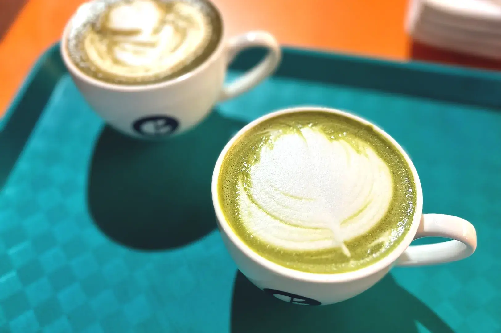 A turquoise tray holding two ceramic cups of matcha latte topped with delicate heart and leaf-shaped latte art.