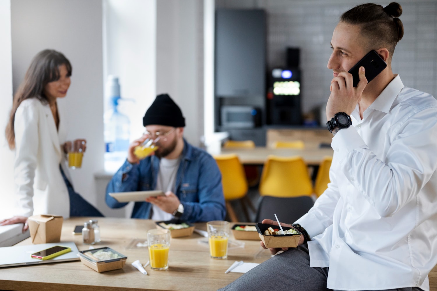 A man sits on a cafeteria table using a smartphone while eating, as two coworkers nearby enjoy lunch and use a tablet. The image reflects modern workplace habits where VoIP phone services and cloud-based phones offer cost-effective communication even during breaks.