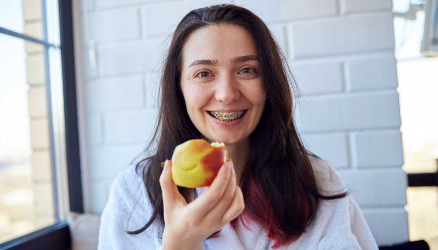 Myk mat ved tannregulering – Beste valg for mindre ubehag 20 Smilende kvinne med regulering holder et eple og ser inn i kameraet.