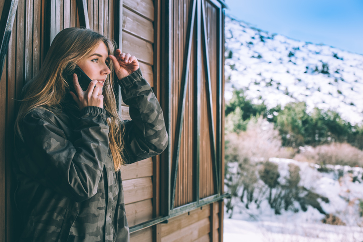Cheerful young woman with long hair in a camo jacket talking on her cell surrounded by mountains.