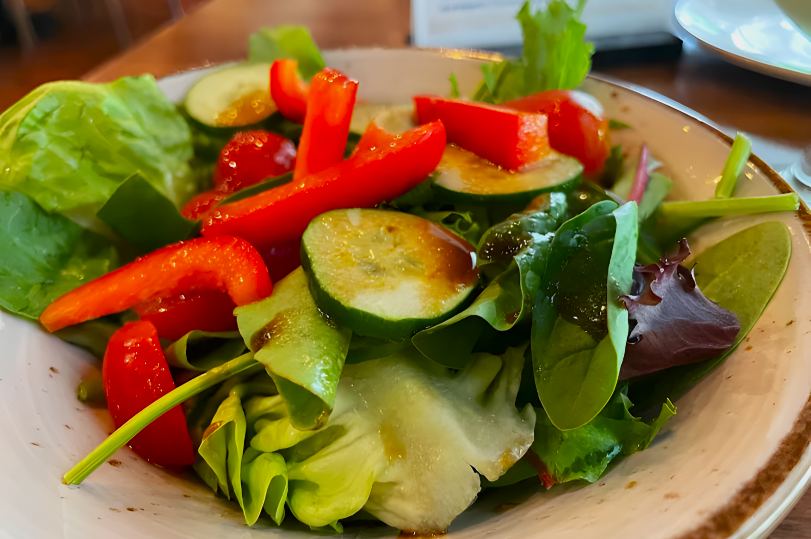 A fresh salad with crisp lettuce, red bell pepper strips, cucumber slices, and cherry tomatoes, drizzled with vinaigrette, served on a white plate.