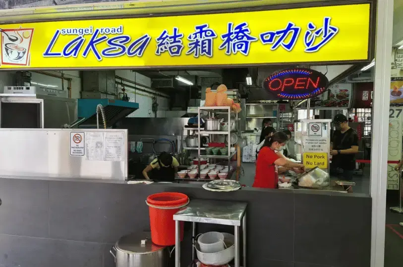A bustling hawker stall named "Sungei Road Laksa" with a bright yellow sign. Inside the compact kitchen, staff members in red and yellow shirts are busy preparing bowls. Stacks of ceramic bowls and a large pot of simmering broth are visible behind the counter.
