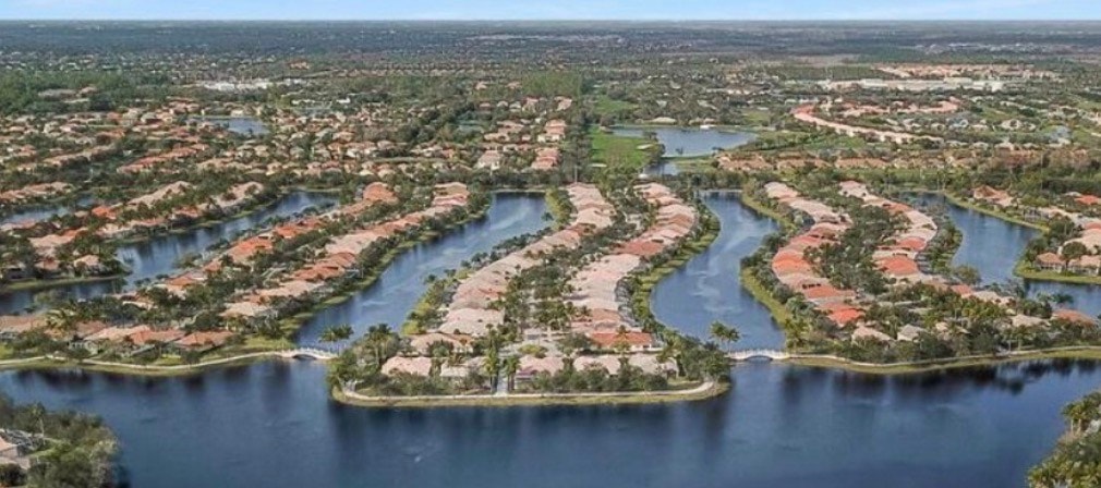 The image depicts an elegant gated entrance to the Island Walk community in Naples, Florida, featuring lush palm landscaping and a secure entry gate. This beautifully maintained area welcomes residents to a desirable community known for its resort-style amenities and vibrant town center complex.