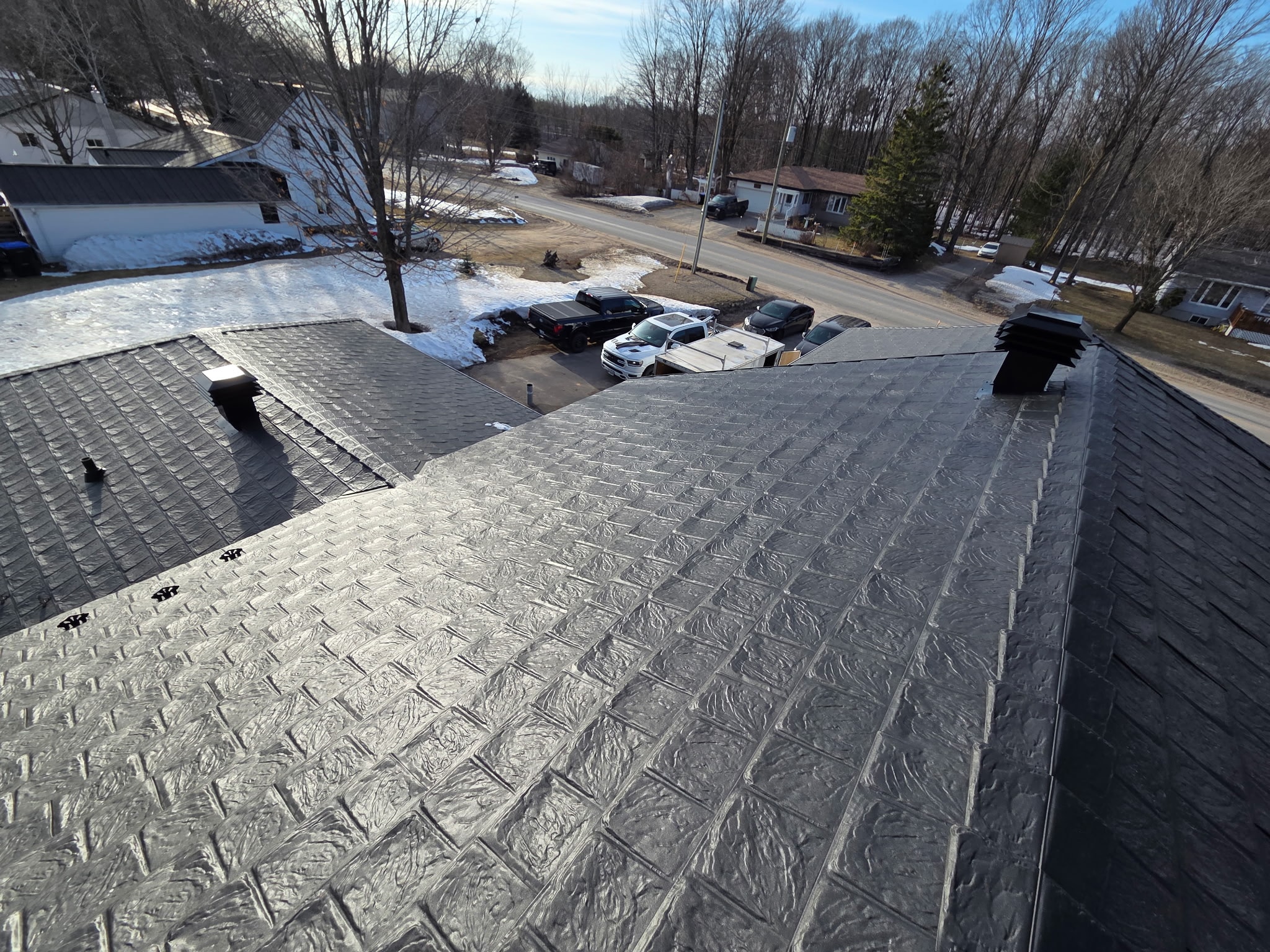 An aerial view of a dark grey metal roof surrounded by vehicles and neighbours. 