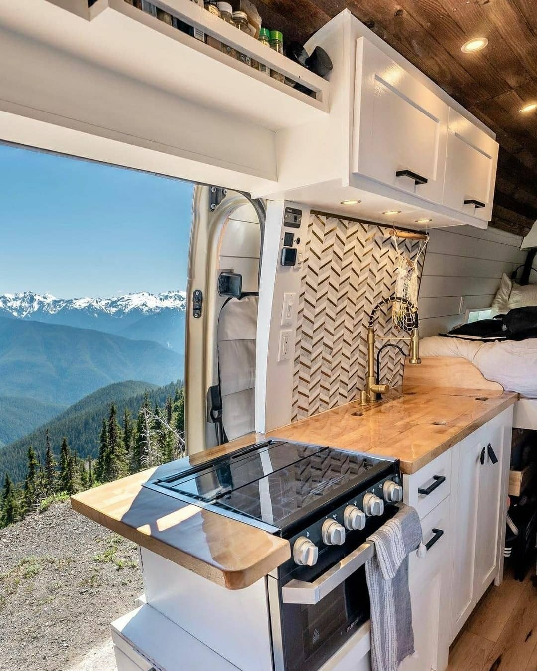 A kitchen counter top with a stove and kitchen faucet within a van. The van door is open displaying an exterior background scene of snow covered mountains. 