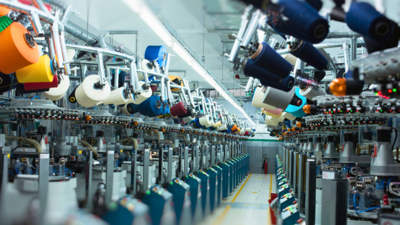 Textile machinery with colorful spools, including orange, yellow, blue, and white, is seen in a bright factory where fabrics are stretched and textured.