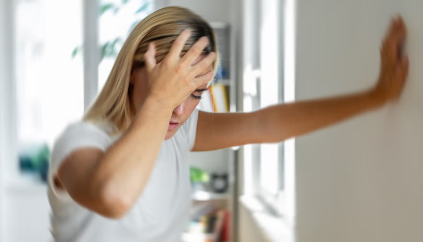 Kvinne holder seg til hodet og støtter seg mot veggen, ser svimmel og utmattet ut i et lyst rom med uklart bakgrunn.