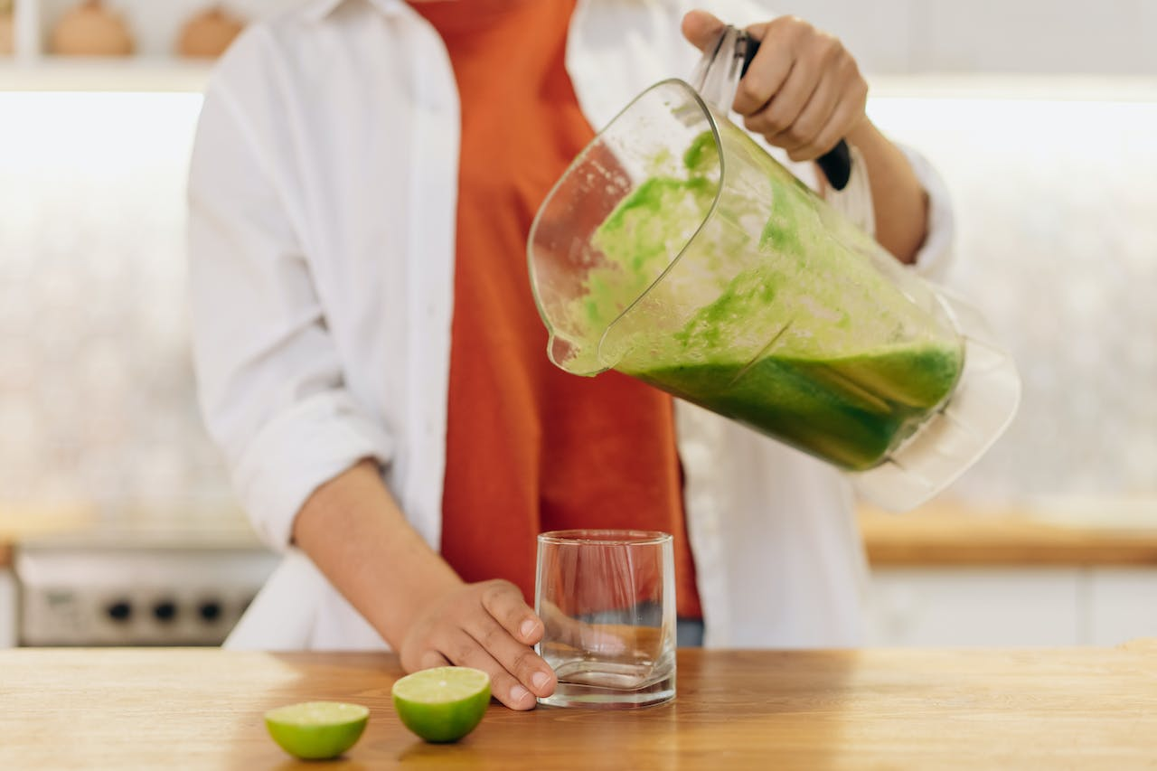 Uma pessoa está vertendo um suco verde de um liquidificador para um copo, com metades de lima ao lado sobre uma superfície de madeira.