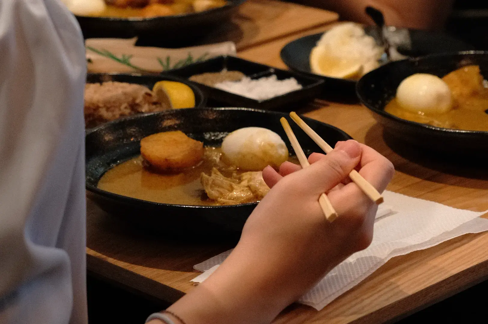A hand holding chopsticks over a bowl of food on a table, with various dishes visible in the background.