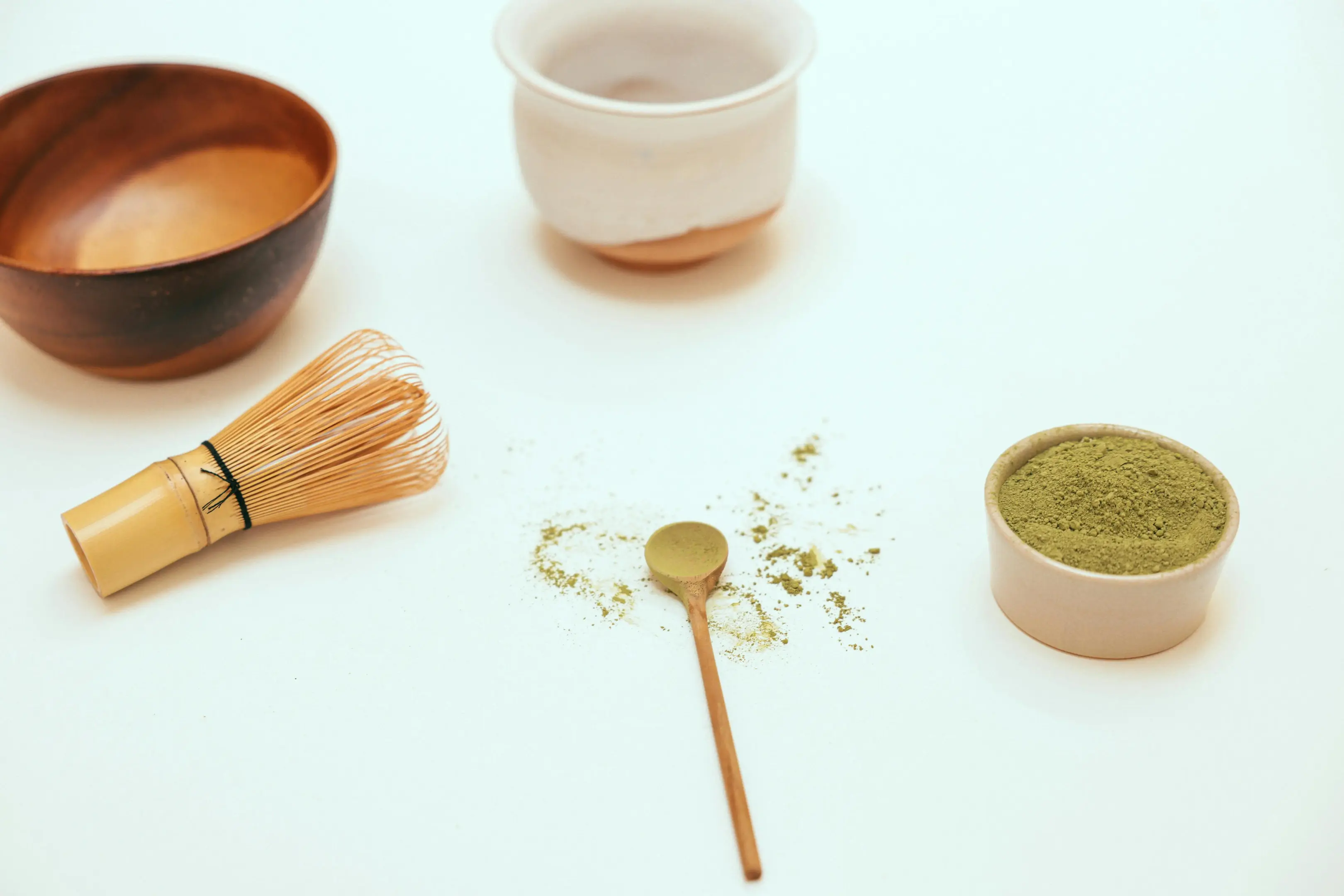 This image shows a minimalist arrangement of matcha preparation tools, including a bamboo whisk, a small wooden spoon, and bowls on a clean white surface. A small container of vibrant green matcha powder sits to the side, with loose powder artfully scattered around the central spoon.