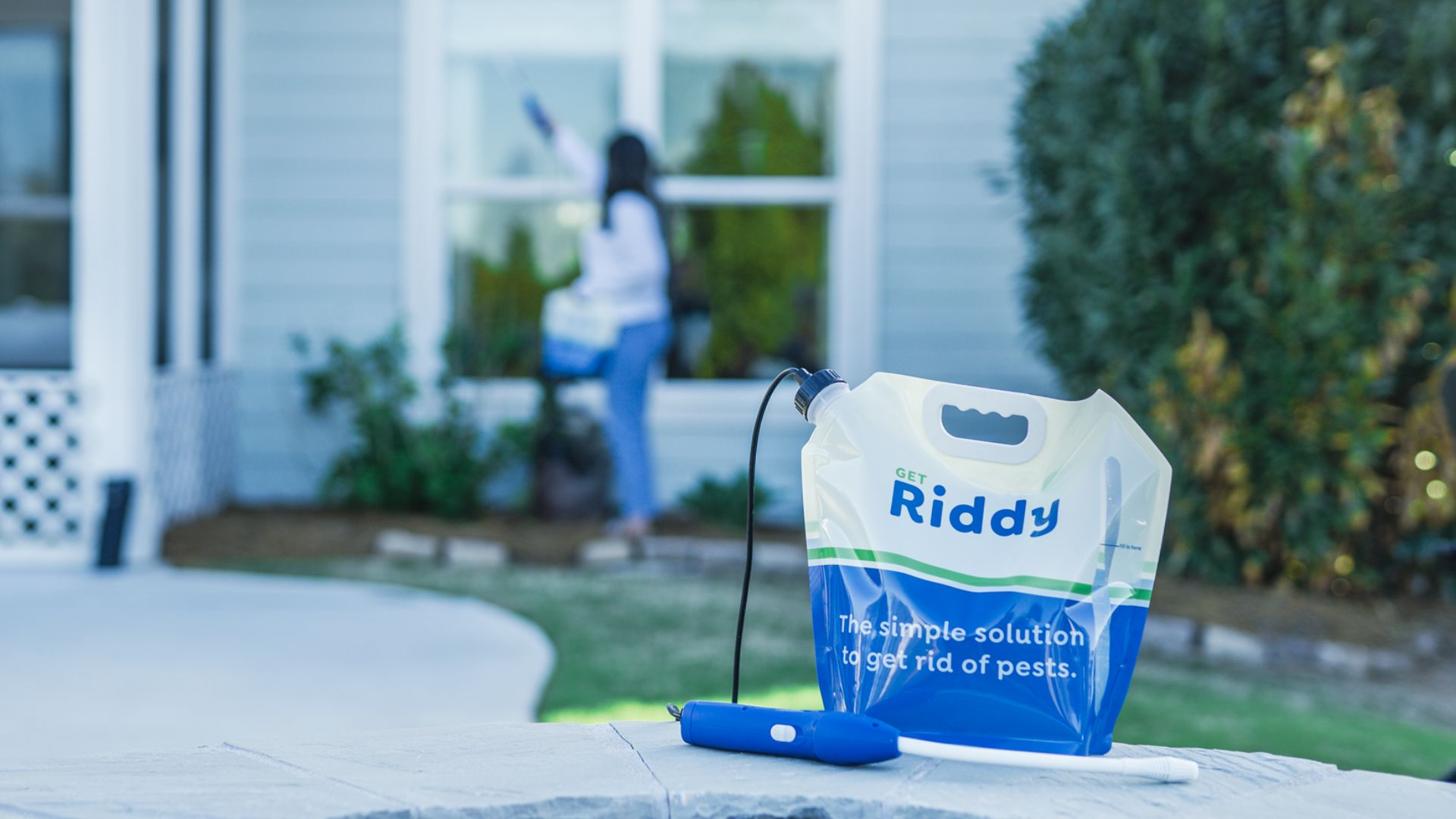 An image of a Riddy insecticide spray bag and applicator wand sitting in front of a home.