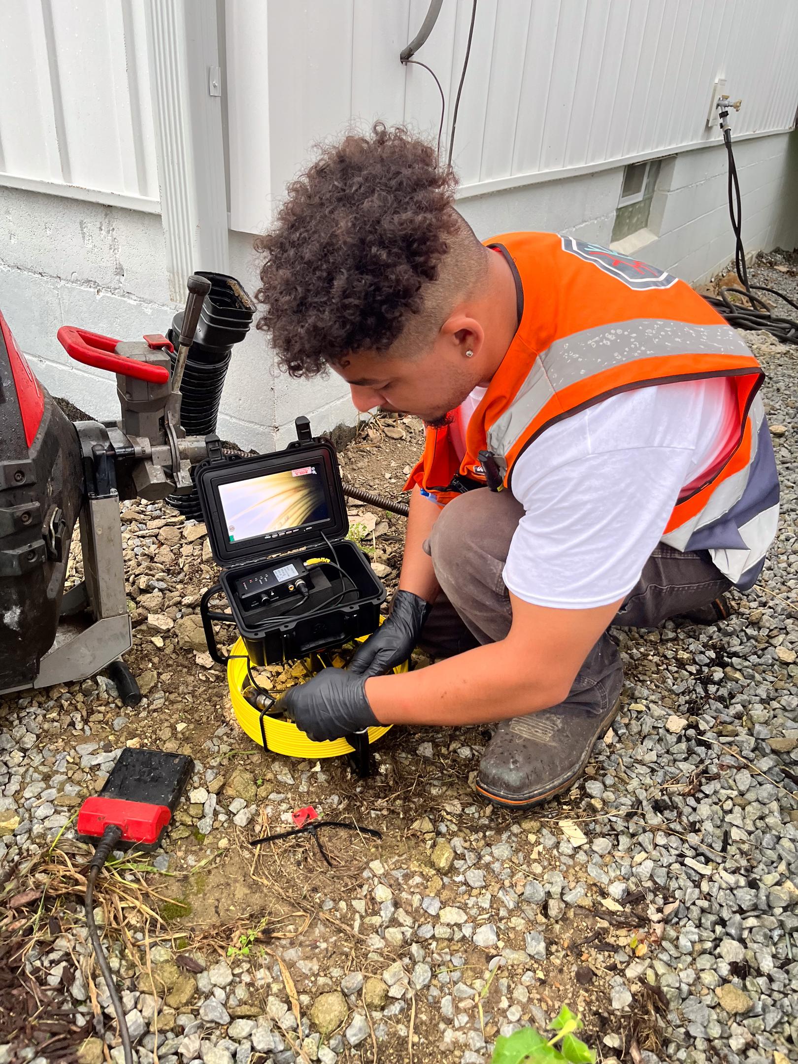 A plumber is examining a residential outdoor cleanout access point, surrounded by various tools and equipment, ready to address potential issues like clogged drains. The scene highlights the importance of regular maintenance in the plumbing system to prevent future problems with water flow and debris buildup.