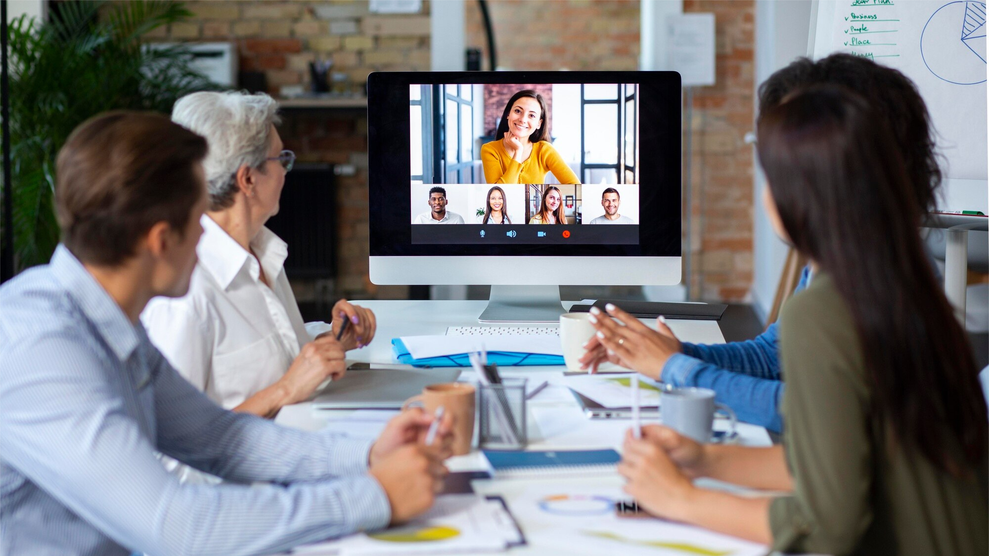 A group of professionals in a modern office gathers around a desk while participating in a hybrid virtual meeting. A large computer monitor shows five remote attendees, demonstrating how video conferencing supports inclusive remote collaboration in business settings.