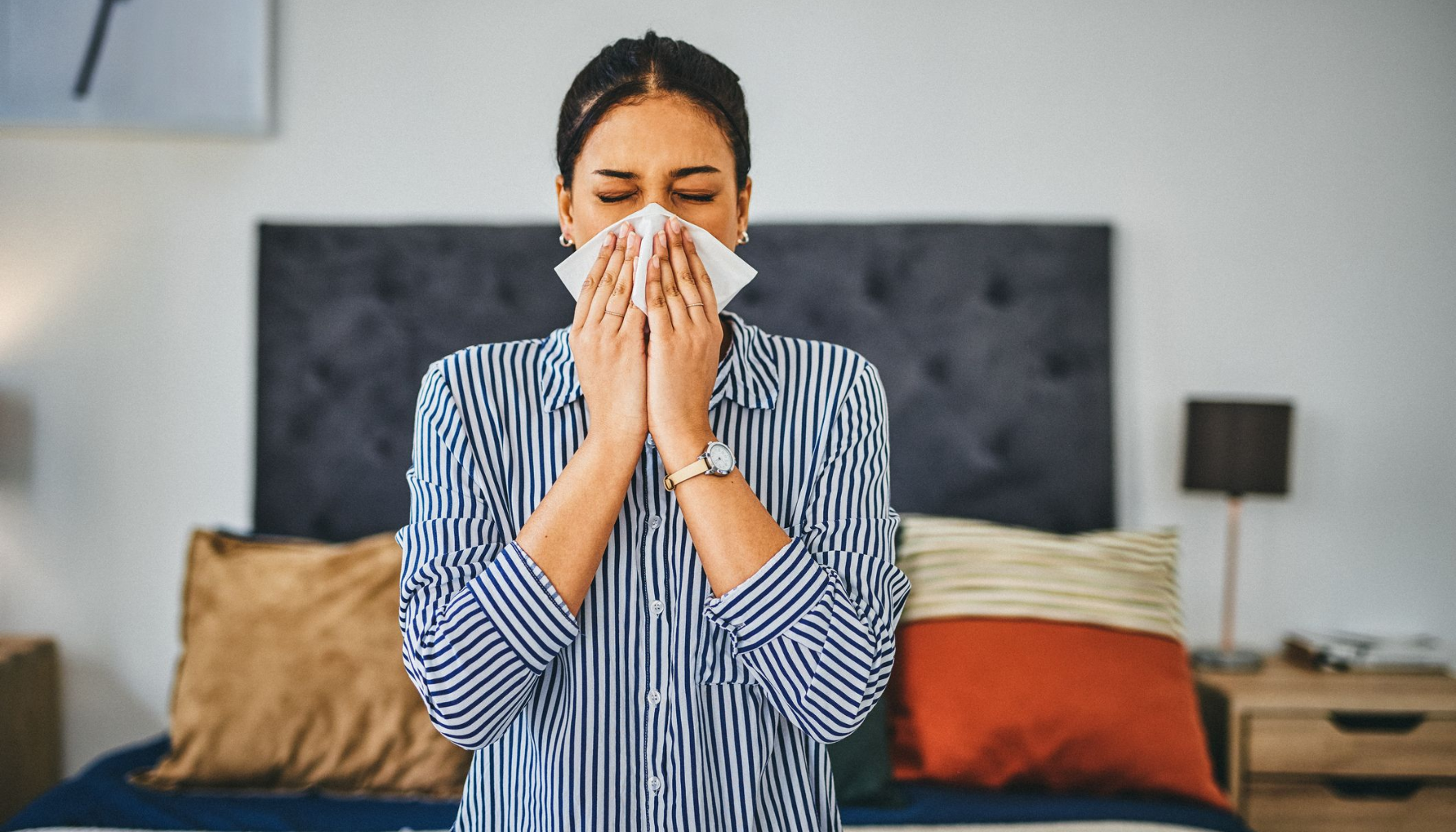 Kvinne i stripete skjorte nyser eller tørker nesen med papirserviett mens hun står ved sengen i et soverom.