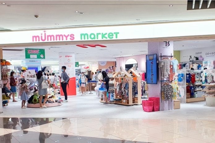 This image shows the storefront of Mummys Market, a retail space filled with a wide variety of baby and children's products. Shoppers, some wearing face masks, browse through organized displays of clothing, toys, and nursery essentials within the bright, modern store.