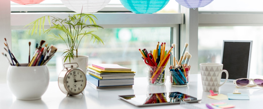 A white desk holding a variety of colourful office and craft supplies including three pen cups, a pile of notebooks, an brass alarm clock, sticky notes and a tablet.