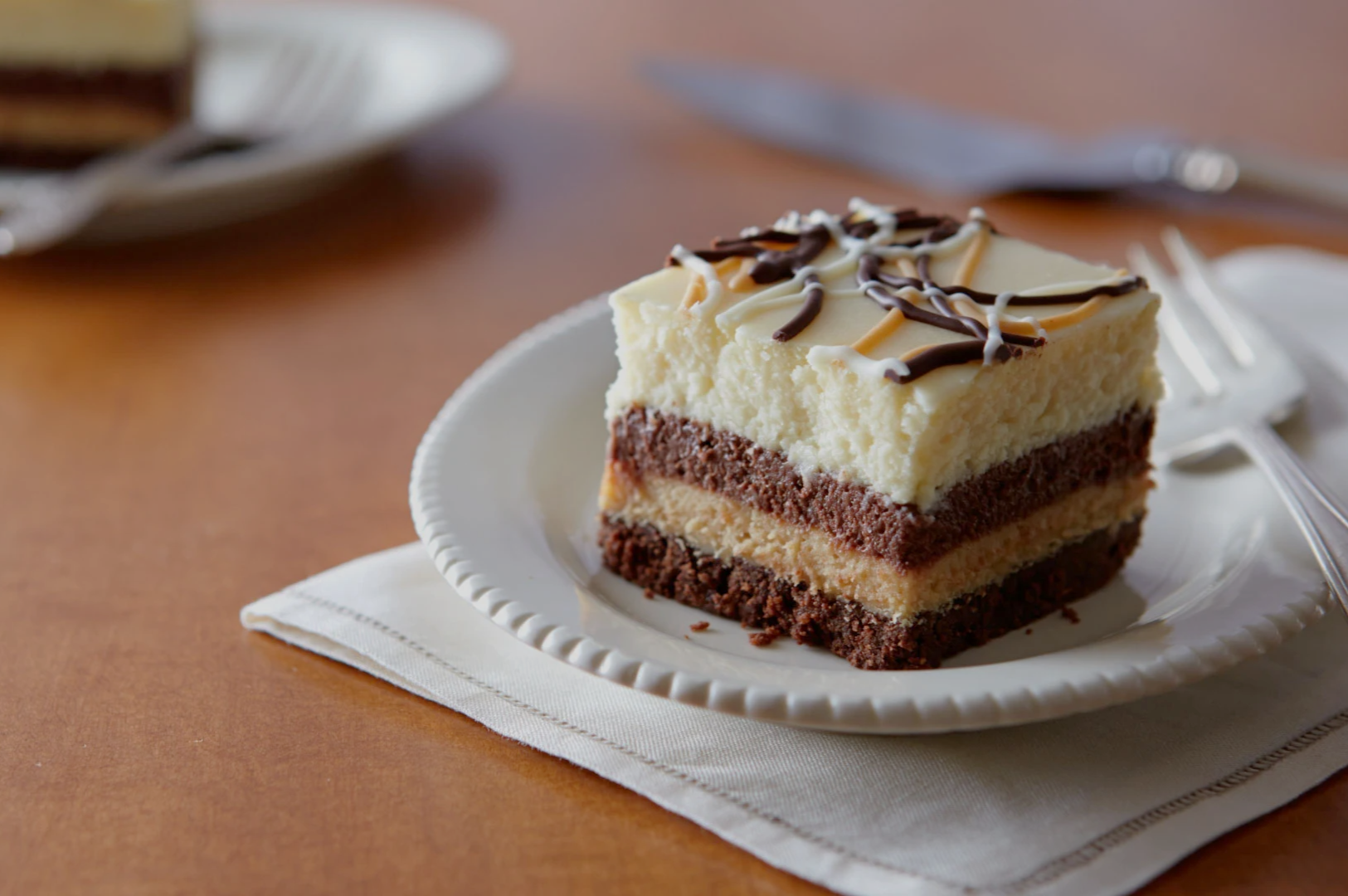 A slice of layered cake with chocolate and cream on a white plate, drizzled with chocolate syrup, placed on a napkin. Background has a knife and another plate.