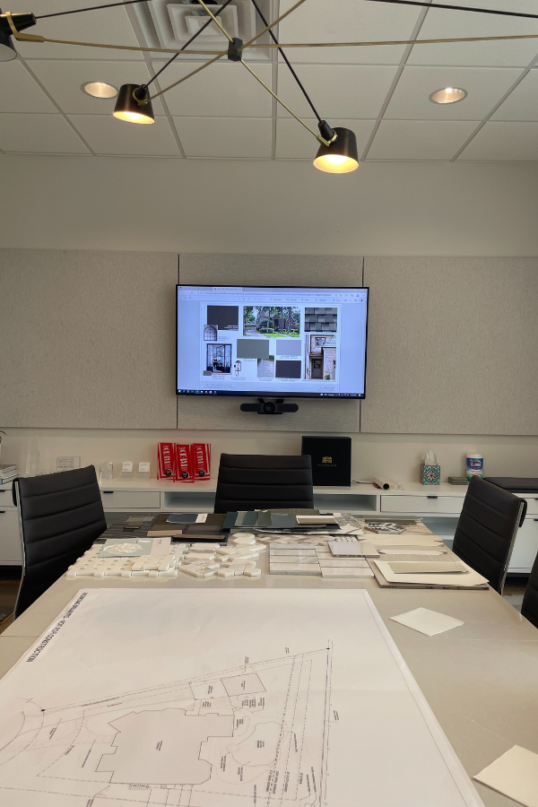 A well-lit design conference room with a table covered in architectural plans, material samples, and design boards; a screen shows reference images on the wall.