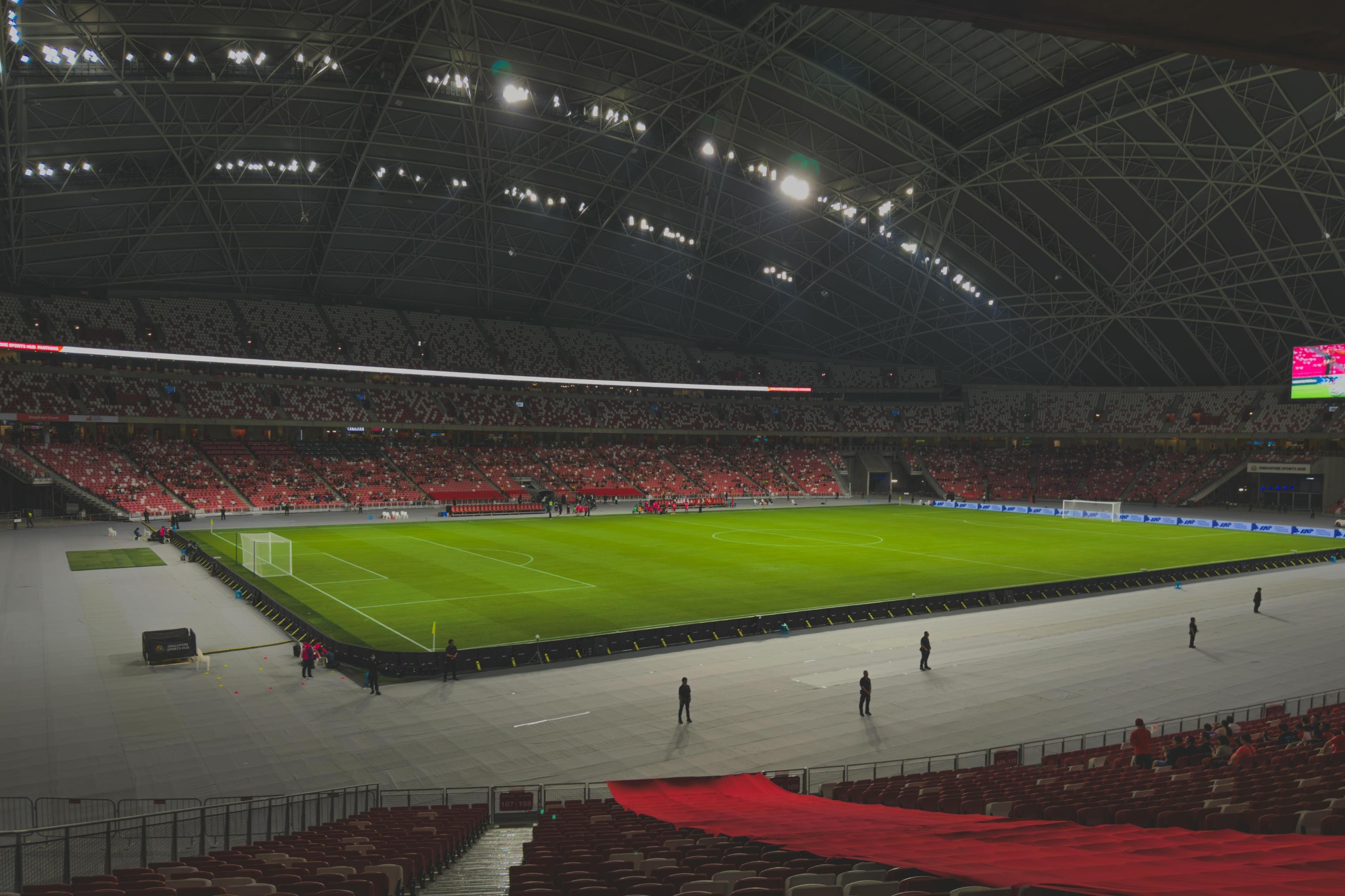 This wide-angle view captures the interior of a massive stadium at night, where bright floodlights illuminate a pristine green football pitch. Tiers of red and white seats rise beneath an intricate metal roof structure, while various figures are scattered along the sidelines of the field.
