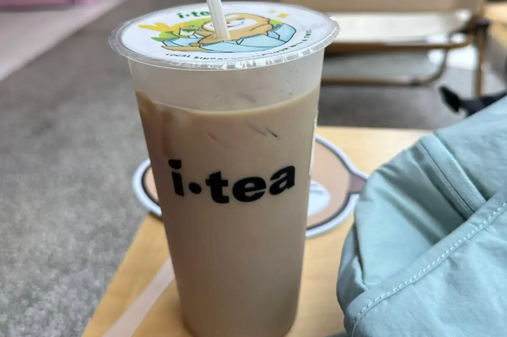 A tall, clear plastic cup of iced milk tea from iTea, sealed with a white plastic film depicting a cartoon otter holding a drink with a straw poked through the top. The cup is held or placed on a light table next to a pale blue fabric bag, showcasing the smooth, light brown milk tea inside.