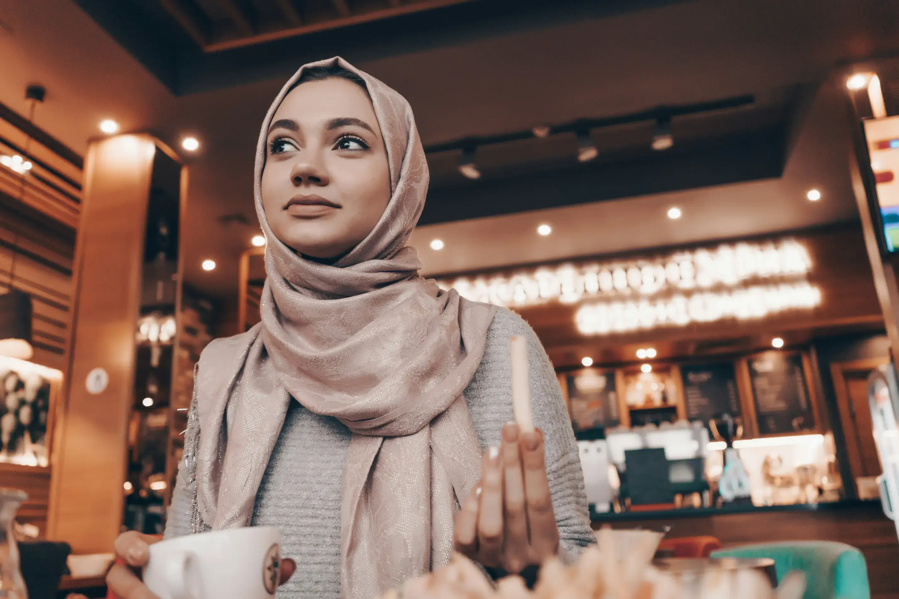 A young woman wearing a shimmering, light pink hijab sits in a cozy café while looking thoughtfully off into the distance. She is holding a white coffee cup in one hand and a french fry in the other, with a blurry background of warm restaurant lights behind her.