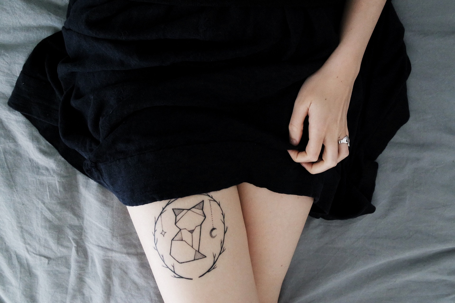 Detalhe mostrando a perna tatuada de uma jovem mulher. A pele é branca e a tatuagem é de um gatinho com formas geométricas, dentro de um círculo feito com pequenos galhos de árvore. A mulher usa um vestido preto rodado e podemos ver parte do seu braço, enquanto ela segura o vestido. Imagem: StockSnap, Pixabay.com.