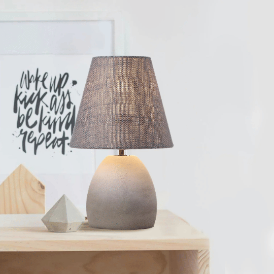 Modern bedside table lamp with a rounded cement base and a dark textured fabric lampshade, placed on a wooden surface next to minimalist decor and wall art.