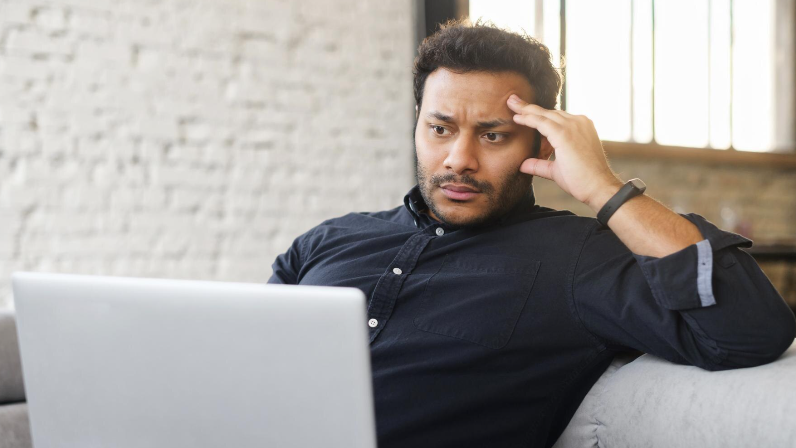 Man sitting on a couch, frowning and concentrating while looking at his laptop, plagued by the common concerns people have about switching to AI-powered VoIP services.