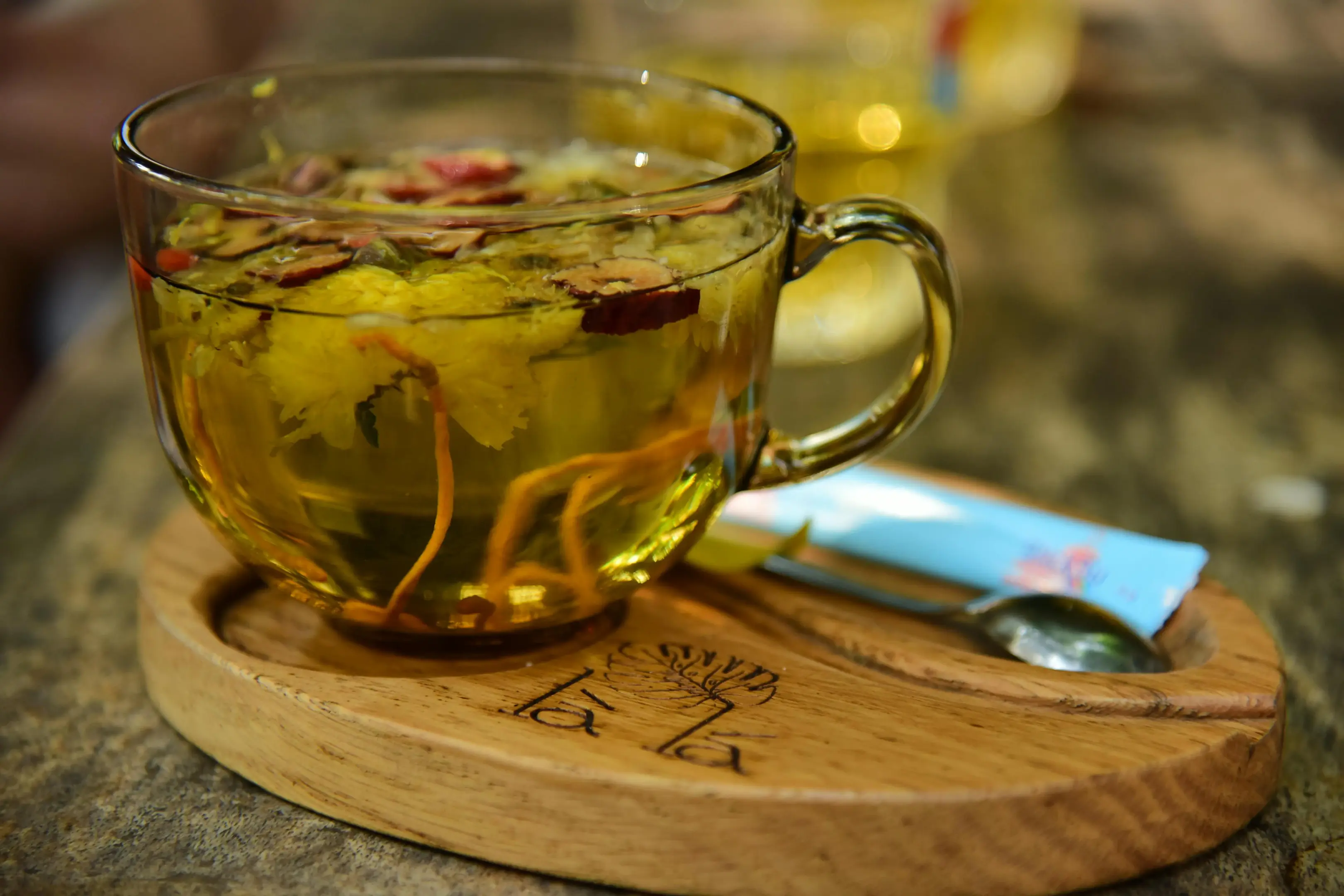 A clear glass mug filled with a vibrant yellow herbal tea sits on a carved wooden saucer, showcasing floating ingredients like chrysanthemum flowers, sliced red dates, and cordyceps. A small metal spoon and a blue packet rest next to the mug on a rustic tabletop.
